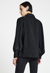 Schwarze Bluse mit langen Ärmeln und lockerem Schnitt, plissierten Schultern und Spitzenverzierung an den Bündchen. Ärmel sind mit Knöpfen abgeschlossen. Strukturiertes Gewebe.