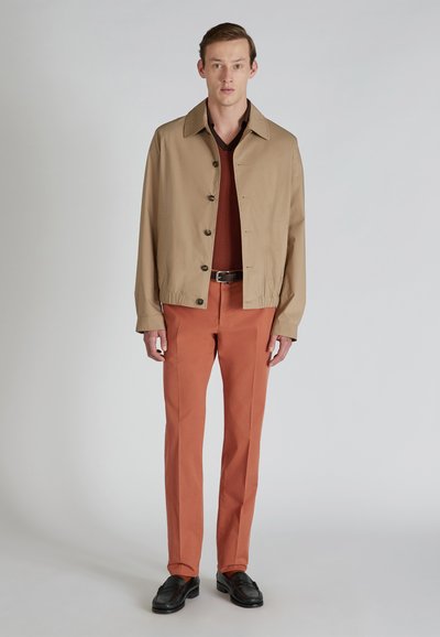 Giacca leggera color sabbia con colletto e chiusura a bottoni frontale, abbinata a un maglione di maglia bordeaux e pantaloni color ruggine. Scarpe nere completano il look.