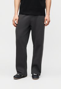 Άνδρας που φορά χαλαρά σκούρα γκρι αθλητικά παντελόνια, μαύρα παπούτσια και μαύρο μπλουζάκι, στέκεται μπροστά σε απλό φωτεινό φόντο.