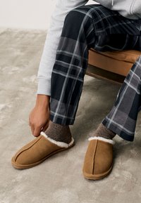 Brązowe zamszowe kapcie z białą podszewką z polaru, o prostym designie i przeszyciu wzdłuż środka, noszone z kratowanymi piżamowymi spodniami.