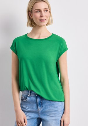 Kobieta z krótkimi blond włosami, ubrana w jasny zielony krótki top i jasnoniebieskie dżinsy, lekko patrząca na lewo na jednolitym tle.