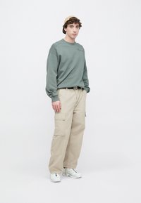 Sudadera verde con cuello redondo, pantalones cargo beige con bolsillos laterales y zapatillas deportivas blancas. El modelo lleva una gorra marrón claro.