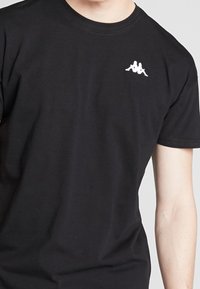 Černé bavlněné tričko s krátkým rukávem. Na levé části hrudi je bílé vyšívané logo. Klasický design s kulatým výstřihem.