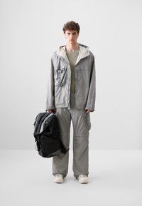 Light gray jacket with white lining and front pocket, paired with matching pants. Holding a black backpack, wearing beige shirt and light shoes.