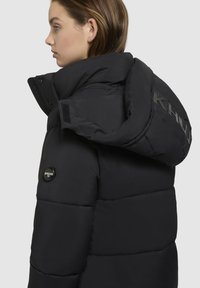 Sort pufferjakke med høj krave, quiltet design og logo-detaljer på siden og huen. Har en sikker lynlåslukning.