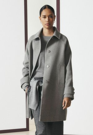 Femme aux cheveux foncés tirés en arrière porte un manteau gris par-dessus un pull gris et un pantalon, se tenant contre un mur blanc uni avec un cadre sombre.