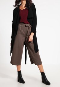 Cardigan long noir ouvert sur le devant, haut bordeaux et pantalon taupe large. Bottines noires complètent la tenue. Texture lisse, coupe décontractée.
