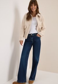 Veste en polaire beige avec fermeture à boutons, deux poches frontales, associée à un jean large bleu foncé et des chaussures marron clair. Design simple.