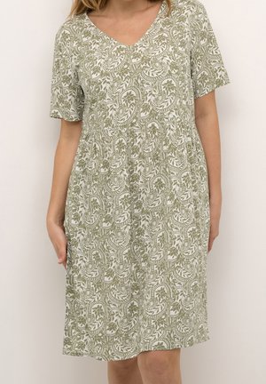 Kvinde iført knælængde kjole med korte ærmer i grøn og hvid blomstret paisley-mønster og V-udskæring mod ensfarvet baggrund.