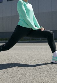 Ljusblå långärmad sporttop som matchas med svarta byxor. Personen sträcker på sig vilket visar en sportig look med gröna detaljer på skorna.