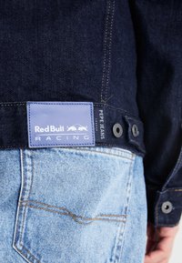 Mörka denimjeans med en ljusblå tvätt längst ner, med en blå läderetikett med "Red Bull Racing" och metallaccenter.