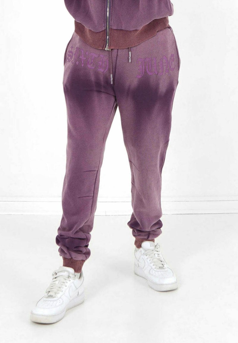 Lila sweatpants med en gradient-effekt, med "SATH" och "JUNE" tryckt i en mörkare lila nyans. Mjuk tyg och elastiska muddar.