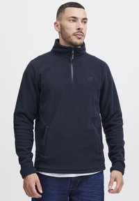 Sudadera de forro polar azul oscuro con un alto cuello y cierre de media cremallera. Presenta bolsillos laterales y un sutil logo en el pecho. Textura suave.