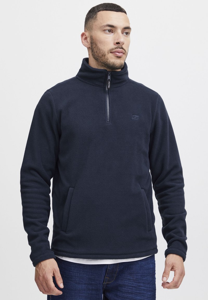 Sudadera de forro polar azul oscuro con un alto cuello y cierre de media cremallera. Presenta bolsillos laterales y un sutil logo en el pecho. Textura suave.