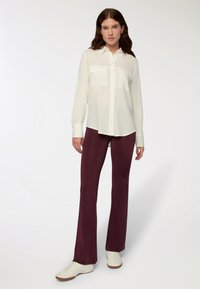 Camicia bianca con bottoni e due tasche sul petto, abbinata a pantaloni a palazzo bordeaux e sneaker bianche, esposta su uno sfondo chiaro.