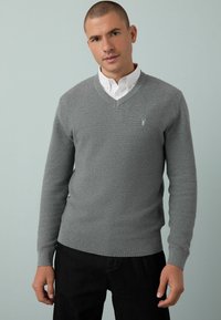 Suéter gris de manga larga con escote en V, de tejido texturizado y puños acanalados. Pequeño logo bordado en el pecho. Lleva puesto sobre una camisa blanca con cuello.