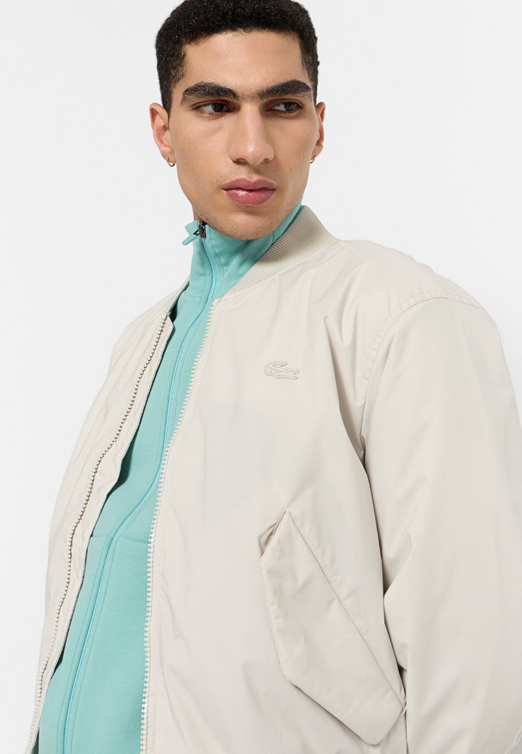 Giacca bomber beige con una texture liscia, dotata di zip frontale e tasca laterale. Indossata sopra una giacca blu con zip. Logo Lacoste presente.