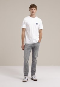 Camiseta de algodón blanca con un logo circular azul en el pecho izquierdo, combinada con jeans gris claro y zapatillas blancas con rayas negras.