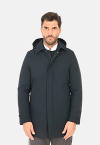 Parka impermeabile blu navy con cappuccio removibile, zip frontale, due tasche laterali e una texture liscia. Presenta un taglio sartoriale e cuciture discrete.