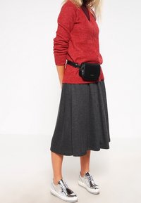 Maglione rosso testurizzato, marsupio nero, gonna plissettata grigia e scarpe con platform argentate creano un outfit stratificato e casual.