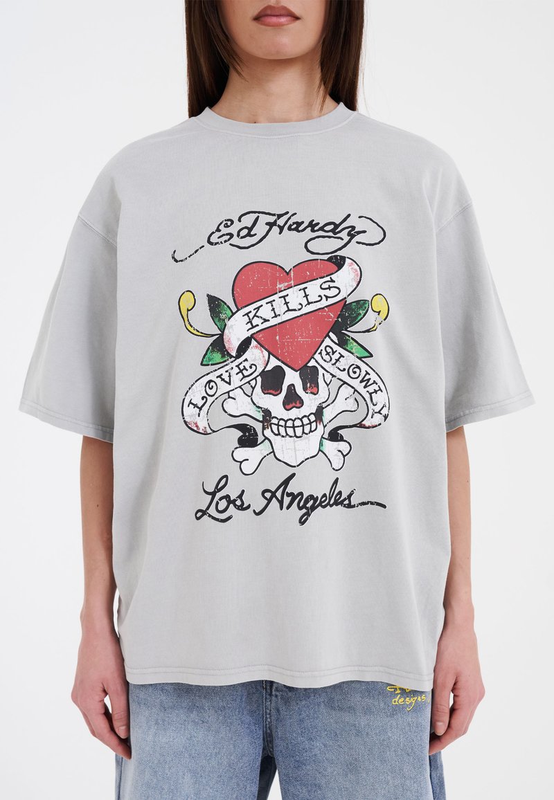 Graues, übergroßes T-Shirt mit einer Grafikdesign von einem Herz, einem Totenkopf und Text in Rot und Schwarz. Aus Baumwollmaterial mit Rundhalsausschnitt.