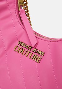 Rosa läderhandväska med en kviltad design, som har en guldfärgad "VERSACE JEANS COUTURE"-logotyp och dragkedja med kedjedetalj.