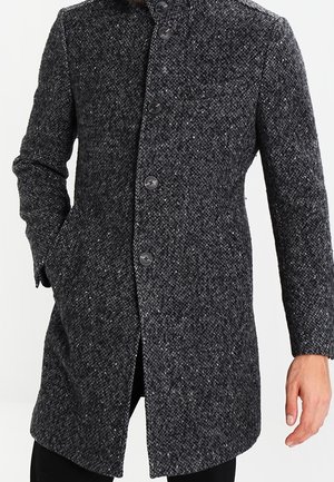 Homme portant un pardessus boutonné gris foncé texturé avec un col montant et une main dans la poche.