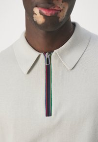 Světle béžová pletená polo košile s límcem na zip a vícolorovými pruhy v červené, zelené a modré barvě podél zipu.