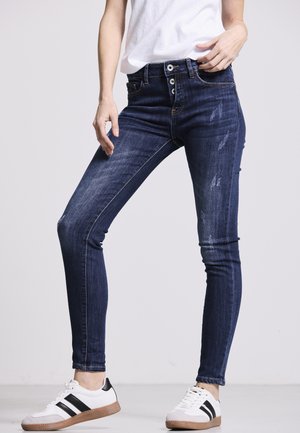 Dunkelblaue Skinny-Jeans mit einem abgenutzten Muster, hoher Taille, fünf Taschen und einem Knopfverschluss, kombiniert mit weißen Turnschuhen.