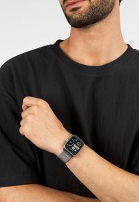 Smartwatch con quadrante rettangolare nero, cinturino in maglia argentata e display digitale che mostra l'ora e le metriche di fitness, indossato su un polso.