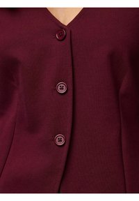 Bordo pleteni kardigan s V-izrezom, četiri dugmeta u istoj boji na prednjoj strani i glatkom teksturom s minimalnim detaljima šavova.