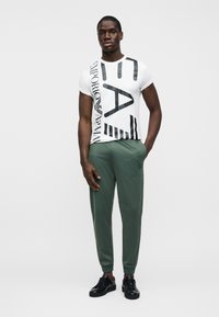 Άσπρο βαμβακερό μπλουζάκι με μαύρα γραφικά κείμενα και ρίγες, συνδυασμένο με πράσινο παντελόνι στενής γραμμής από απαλό, ελαφρύ ύφασμα. Μαύρα αθλητικά παπούτσια.