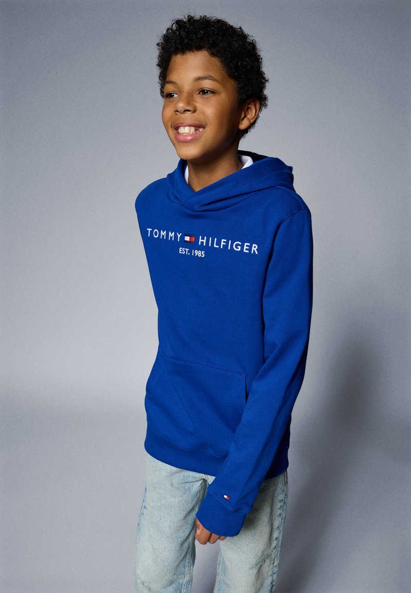 Blå huvtröja med dragsko, som har "TOMMY HILFIGER"-logotyp, en framficka och ribbad mudd. Matchad med ljusblå denimjeans.