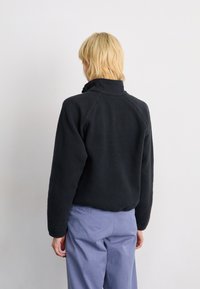Black fleece jacket with a high collar and raglan sleeves, featuring a textured finish. Worn with light blue trousers.