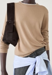 Personne portant un pull couleur chameau, un pantalon gris, une chemise bleu clair nouée à la taille et un sac à bandoulière en daim marron foncé porté en travers de la poitrine.