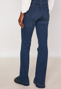 Donna che indossa jeans a vita alta blu scuro con orli sfrangiati, una maglietta bianca a maniche lunghe e scarpe scure, in piedi di fronte a uno sfondo neutro.