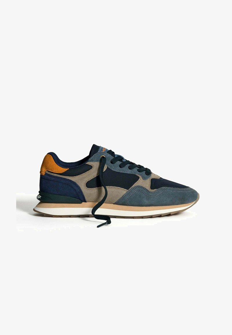 Avslappnad sneakers i marinblått, grått, beige och orange med mockadetaljer, svarta snören och en vit och brun gummisula, fotograferad på vit bakgrund.