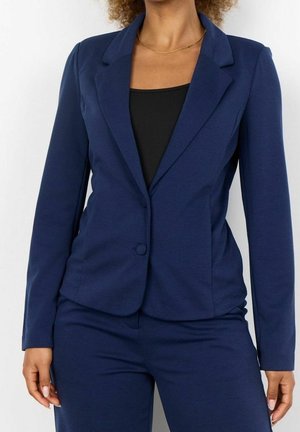 Femme portant un blazer bleu marine ajusté à deux boutons sur un haut noir et un pantalon bleu marine assorti, debout devant un fond blanc.
