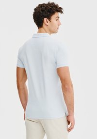 Jasnoniebieska koszulka polo z krótkimi rękawami, kołnierzykiem i bocznymi rozcięciami. Miękka tkanina o gładkiej strukturze, dopasowany krój. Pokazany widok z tyłu.