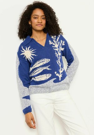 Frau mit lockigem Haar, die einen blauen und grauen Pullover mit Mustern von Fischen, Sonne und Seetang trägt, kombiniert mit weißen Hosen, stehend vor weißem Hintergrund.