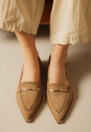 Nohy v béžových semišových špicatých mokasínach s dekoratívnymi perforáciami a zlatými kovovými akcentmi, v kombinácii so svetlými voľnými nohavicami.