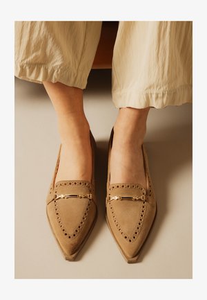 Nohy v béžových semišových špičatých mokasínách s dekorativními perforacemi a zlatými kovovými akcenty, doplněné světle barevnými volnými kalhotami.