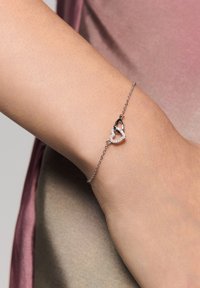 Srebrna bransoletka łańcuszkowa z wzorem serca, jedna połowa w czerni z kryształami, a druga połowa w srebrnym konturze, na tle skóry.