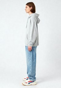 Felpa con cappuccio grigia in cotone con tasca frontale, abbinata a jeans in denim blu chiaro e sneakers bianche con dettagli rossi e blu.