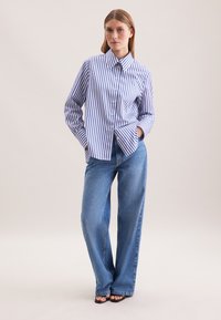 Vrouw staat met handen in de zakken, gekleed in een blauw-wit gestreepte button-up shirt, blauwe wide-leg jeans en zwarte openhaal schoenen.
