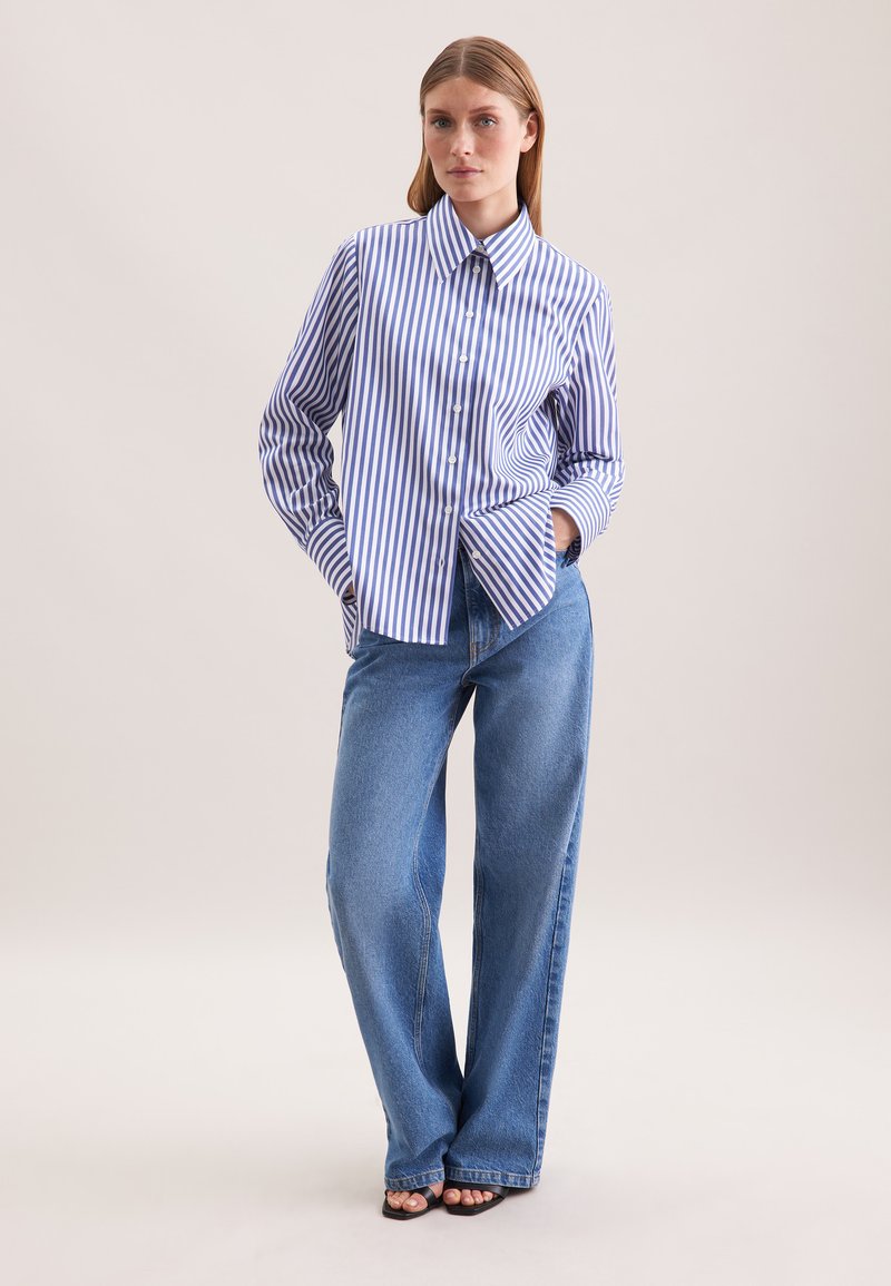 Vrouw staat met handen in de zakken, gekleed in een blauw-wit gestreepte button-up shirt, blauwe wide-leg jeans en zwarte openhaal schoenen.
