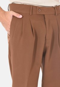Pantaloni a pieghe marroni realizzati in tessuto morbido con chiusura a bottone, dotati di tasca laterale e texture sottile. Vestibilità tailored, design moderno.