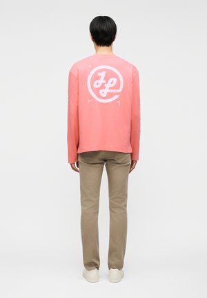 Inimene, kes kannab korallivärvi pikkade varrukatega särki valge ringikujulise "JL" logoga seljal, beežid pükse ja valgeid tossusid, seisab selg vaataja poole.