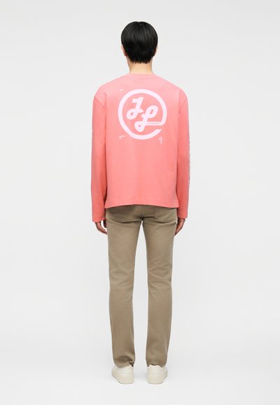 Personne portant une chemise corail à manches longues avec un logo circulaire blanc "JL" au dos, un pantalon beige et des baskets blanches, debout de dos.