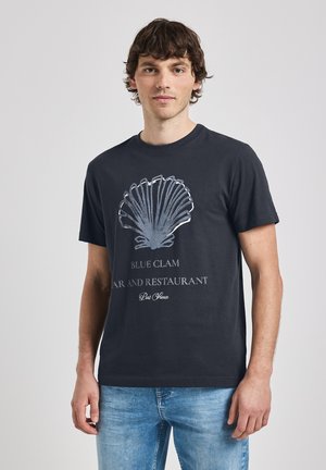 T-shirt en coton bleu marine foncé avec un grand graphique de coquille argentée et le texte "BLUE CLAM BAR AND RESTAURANT" en blanc et gris clair.